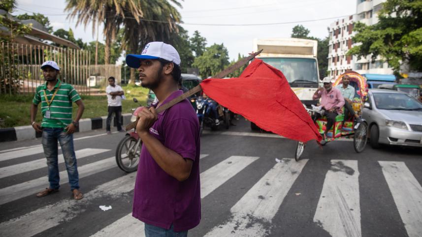 A student managing the traffic at Bijoy Sarani while resting a red flag on his shoulder.  